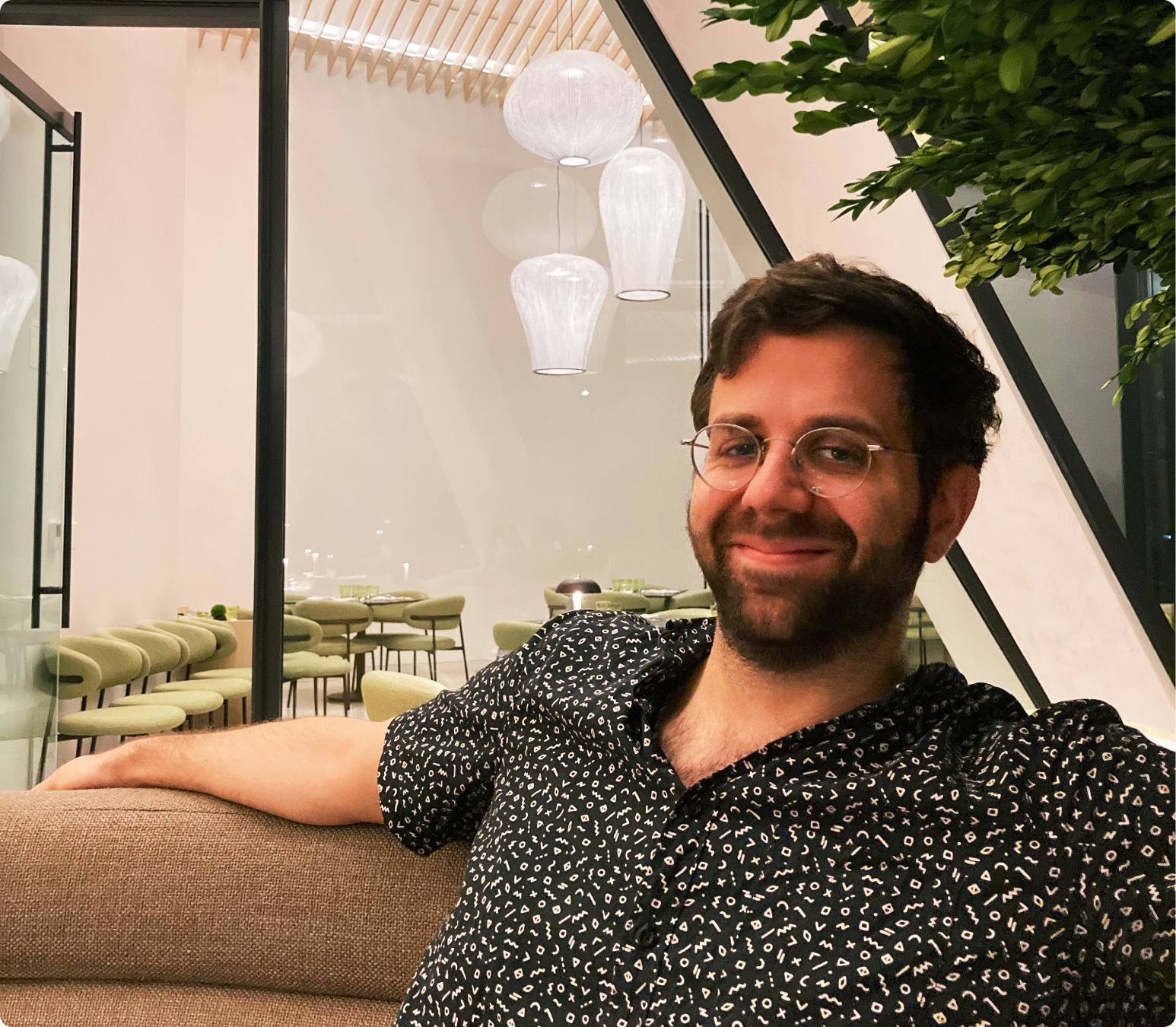 Corbin Lawrence smiling, seated on a couch in a plant-filled modern restaurant